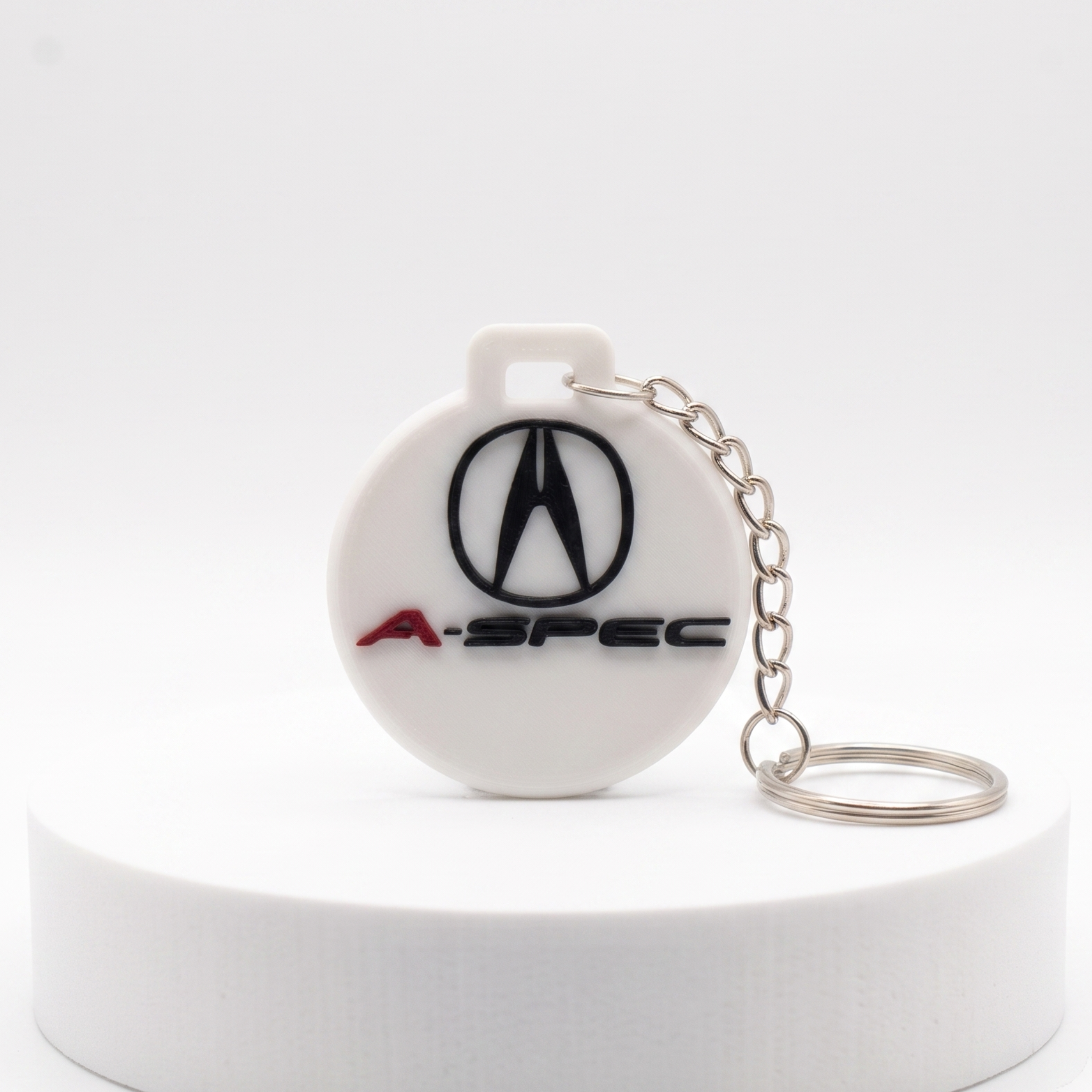 A white circular A-Spec keychain featuring the black Acura logo and red A-SPEC text, attached to a silver metal chain, displayed on a white cylindrical pedestal against a white background.