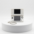 A miniature white handheld game console keychain replica with a black screen and silver keyring, displayed on a white cylindrical pedestal against a white background.