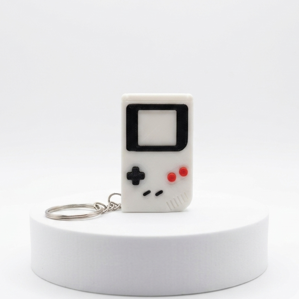 A retro handheld game console keychain with a white body, black D-pad and screen outline, and red buttons, displayed on a white cylindrical pedestal against a white background.