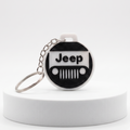 A circular keychain featuring a black and white Jeep grille design with the word 'Jeep' on the front, attached to a silver metal key ring and displayed on a white cylindrical pedestal against a light gray background.