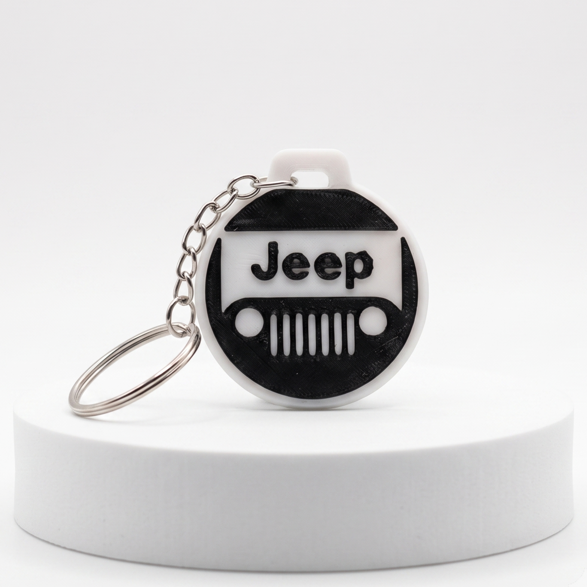 A circular keychain featuring a black and white Jeep grille design with the word 'Jeep' on the front, attached to a silver metal key ring and displayed on a white cylindrical pedestal against a light gray background.