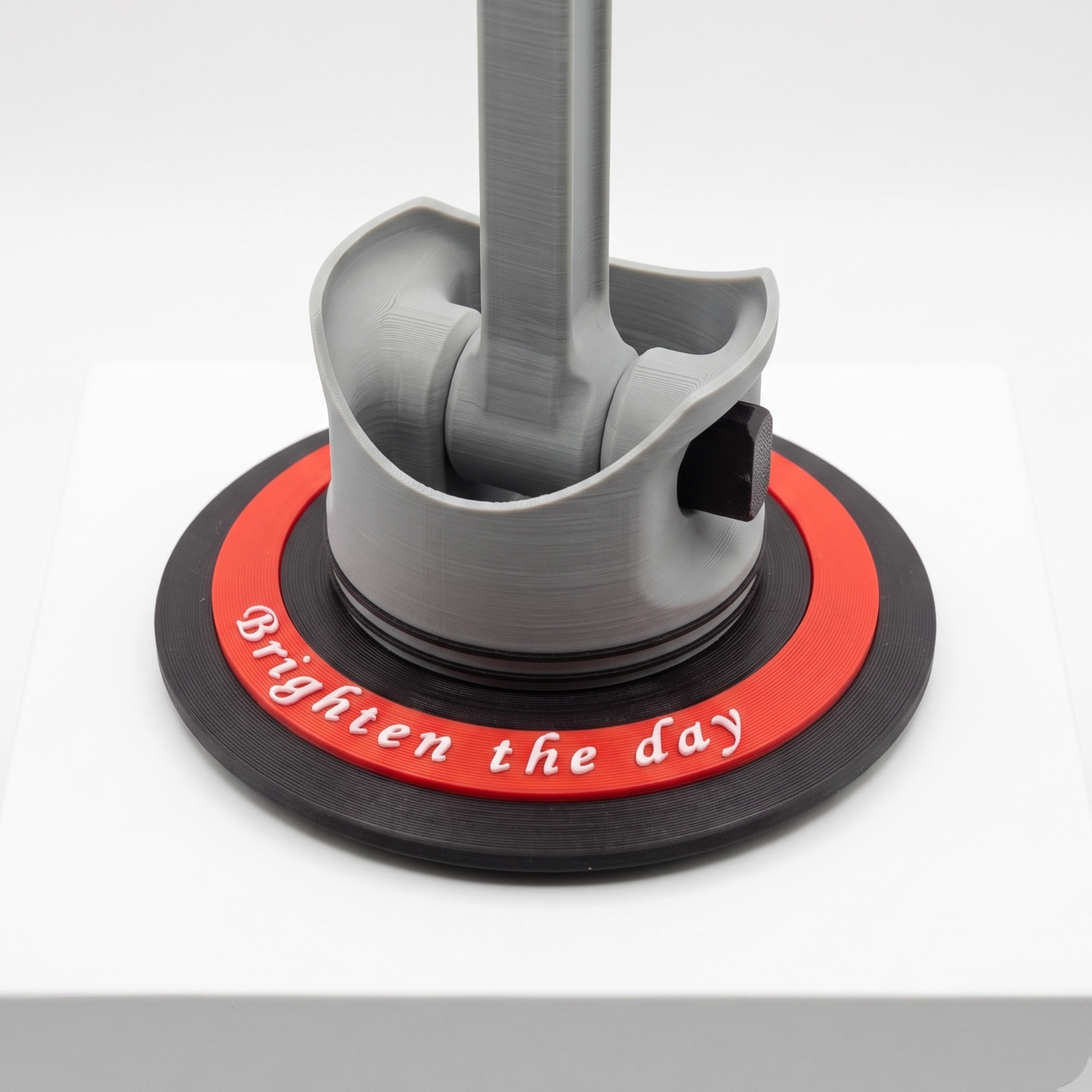 A 3D-printed desk lamp base with a gray metal pole, featuring a black circular base with a red ring and the white text 'Brighten the day'. The lamp is displayed on a white cube against a light gray background.
