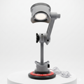 A gray adjustable desk magnifier lamp with a circular head, mounted on a stand with a red circular base, shown on a white cylindrical pedestal against a light gray background.