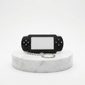 A miniature black handheld gaming console keychain with a silver split ring, displayed on a round white marble pedestal against a clean white background.