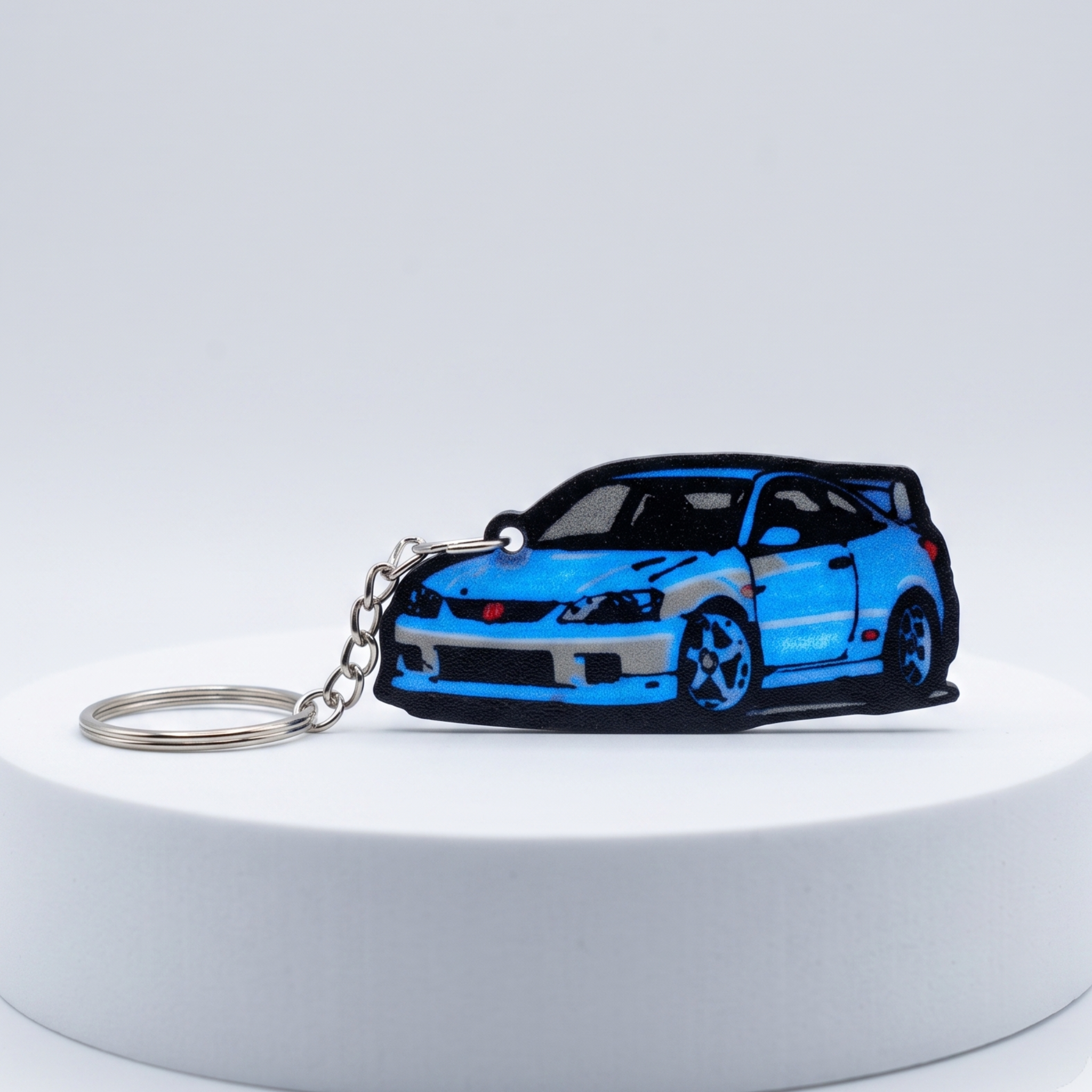 A blue and black car-shaped keychain with a silver metal chain, displayed on a white cylindrical pedestal against a light gray studio background.