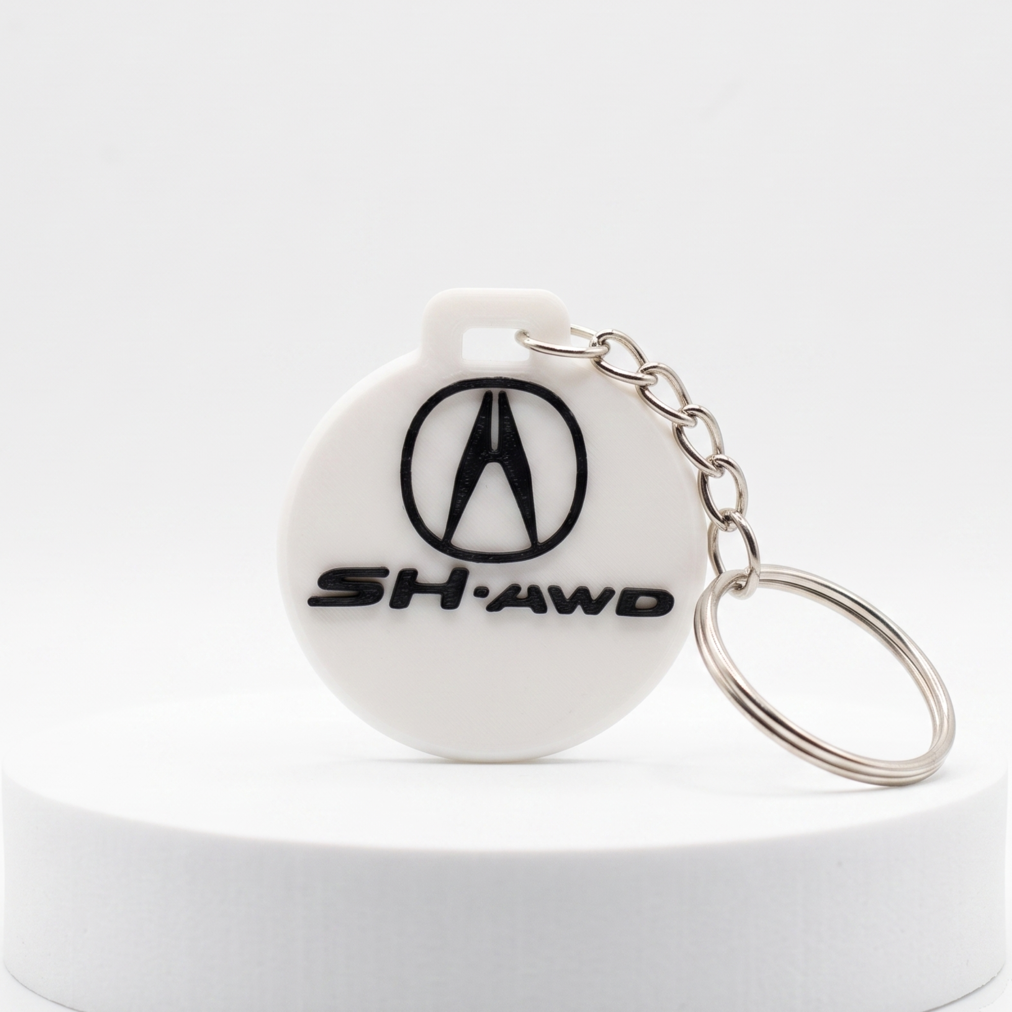 A white circular keychain featuring the Acura logo and 'SH-AWD' text in black, attached to a silver metal key ring, displayed on a white cylindrical pedestal against a white background.