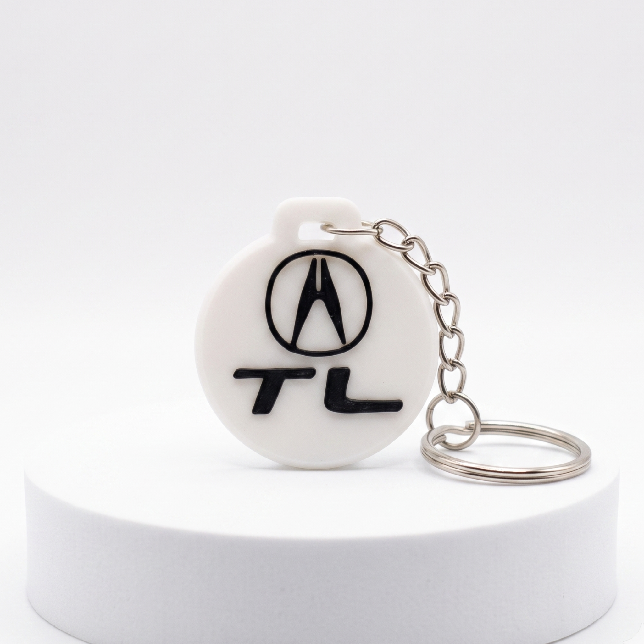 A white round keychain featuring the black Acura logo and 'TL' text, attached to a silver metal chain, displayed on a white cylindrical pedestal against a light gray background.