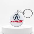 A white circular keychain featuring the Acura logo and 'TYPE S' text, attached to a silver split ring and displayed on a white pedestal.